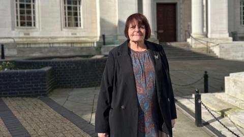 Hazel Simmons stands on a paved walkway in front of a large stone building with tall columns and symmetrical windows. The person is wearing a patterned dress with a dark jacket, and sunlight casts strong shadows across the scene.