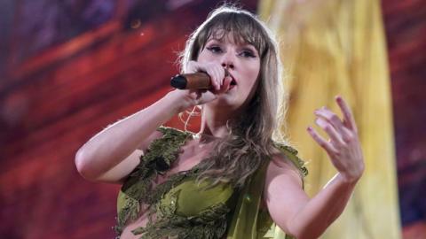 Taylor Swift wearing a green dress and holding a microphone during an Eras tour performance in Munich