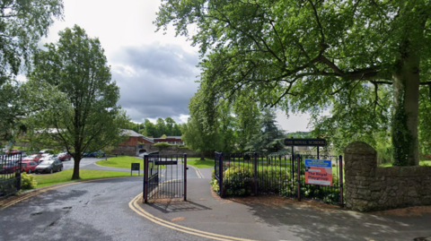 A screengrab of a Google Maps image of the tree-lined entrance to the Royal School, Armagh on a sunny day. A stone exterior wall is partially visible and black metal railings line separate pedestrian and vehicular entrance to the site. Behind the railings is a black sign which says "The Royal School" in gold lettering. There is a carpark and a modern red-brick building in the background.