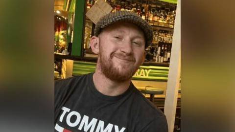 Family photograph of Patrick Shaughnessy. He is wearing a peak cap and has a ginger beard and moustache. He is standing in a bar with lots of bottles on a shelf in the background.