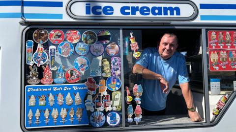 Mark Edwards looking out of the window of his ice cream van. He is wearing a blue t shirt and is looking at the camera.