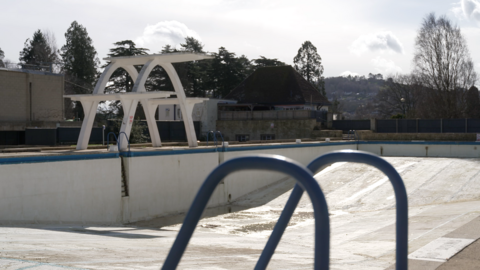 An wide vista of a large, empty open swimming pool with a white two-stage parabolic diving board in the background. 