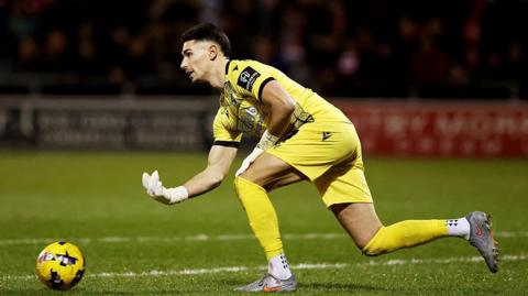 Luke Southwood, wearing an all-yellow kit, bowls the ball out along the ground for Bristol Rovers