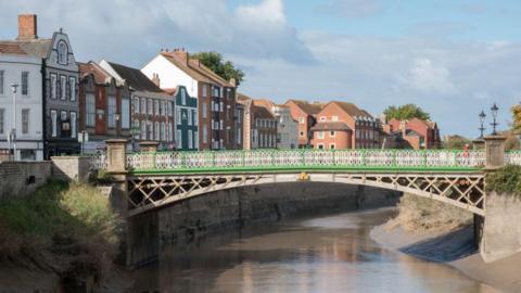 A bridge is connecting two roads over a river in Bridgwater. To the left of the river are town houses. A person is crossing the bridge, which has green and white railings.