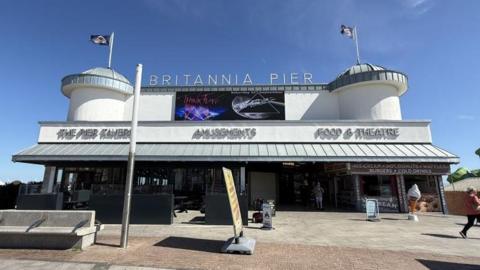 A front view of Britannia Pier that has flags flying from both its two turrets. The building is grey and cream and has letters standing on its top saying: Britannia Pier. Below this sign are other signs that say: The Pier Tavern, Amusements and Food & Theatre. The sky is blue.
