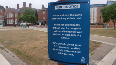 Blue sign tied to a lampost apologising for poor state of grass
