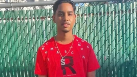 Shawn Seesahai standing in front of a green gate with a red t shirt and a necklace with an S pendant