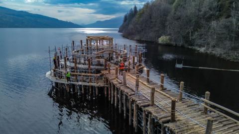 A recreation of an Iron Age roundhouse under construction set on a loch