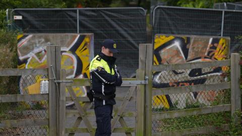 A man wears a black and hi-viz yellow jacket with black trousers. He wears a navy baseball cap. Behind him is a fence with a black sheet on it. protecting an area of green woodland.