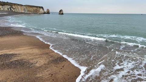 Photo taken from a sandy beach near to where the water is lapping on to the coast. Cliffs are in the distance, breaking off into columns of rock.