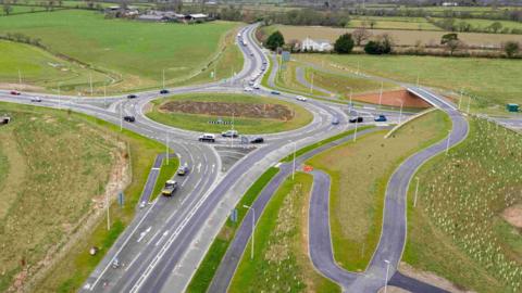 An aerial view of a roundabout with new linking roads around it. One is going over a bridge over the top of one road feeding into the roundabout. Tree saplings have been planted on the banks around the roundabout.
