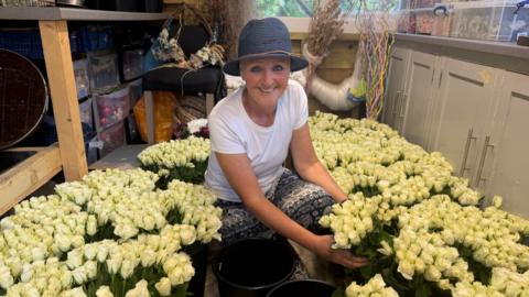 Karen Simpkin in a shed crouched down, surrounded by bouquets of white roses.