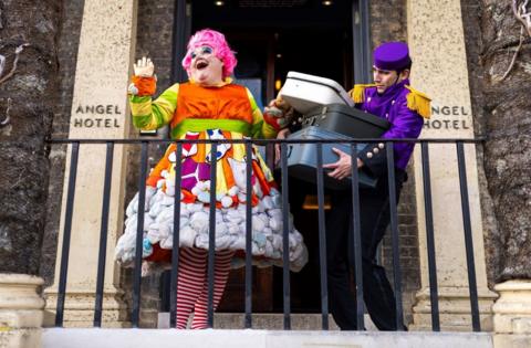 A panto dame and a hotel bell boy stand on the front steps of the Angel Hotel in Bury St Edmunds. The dame is wearing a colourful outfit with a pink wig, while the bell boy has a purple uniform on. They are characters from Aladdin which is running at the Theatre Royal in the town