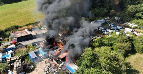 A close up drone view of a scrapyard fire, surrounded by green fields, with a number of shipping containers on fire and thick black smoke rising