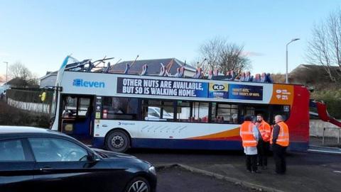 A white bus with red and orange and a blue advert on the side without a roof. The roof is hanging off the back of the vehicle.