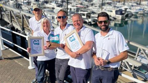 A picture of five people stood leaning on railings in front of the marina.  They are all wearing white polo shirts with a number of people holding framed awards.