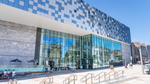 The exterior of The Box museum and art gallery. The ground floor has large glass panel windows. There are people sat outside of the museum and people walking to the right. There is a blue square design above the windows.