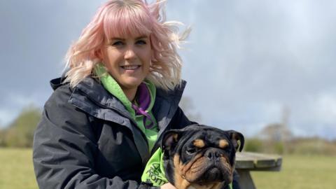 A woman with blonde and pink hair sits on a bench holding a small black dog.