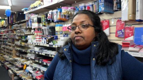 A woman wearing a dark blue gilet and turtle-neck jumper is standing inside a shop.