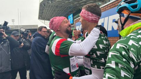 Two men in a green, white and red rugby tops embrace at a rugby ground as photographers take photos. 