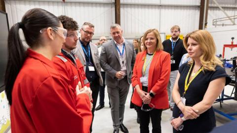 North East mayor Kim McGuinness and North Tyneside mayor Karen Clark visiting the Newcastle College Energy Academy in Wallsend. They are standing in a large room and talking to two students dressed in red overalls and wearing safety goggles, while other people watch them.