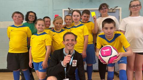A young man dressed in black and holding a whistle kneels down in a sports hall. He is surrounded by young children in matching yellow sports kits holding footballs