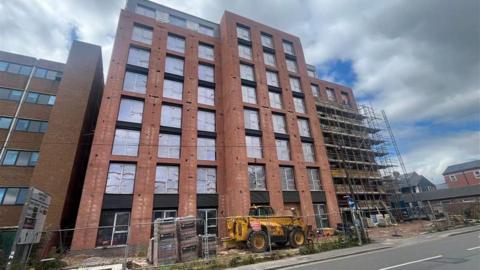 Image of a red-brick eight-storey building site with a digger in the foreground