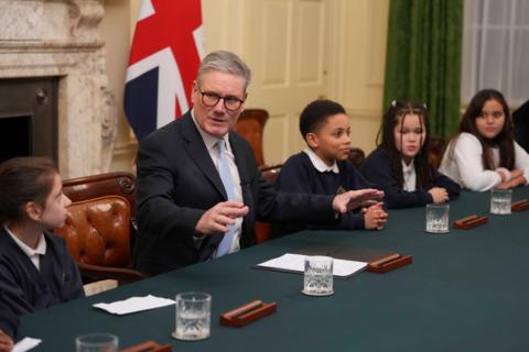 prime minister keir starmer speaking to children in the cabinet room of number 10 with everyone sat at a table with glasses of water. the flag of the united kingdom stands behind him and the students.