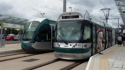 Two green Nottingham tram at in the day stationary at one of the stops. The image includes people alighting one of the trams.