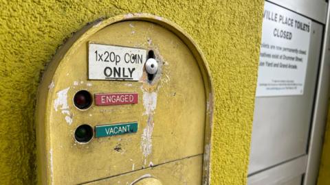 Closed public toilets. There is a "1x20p Coin Only" sign which also says "Engaged" and "Vacant".