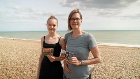 Two women pose holding postcards on Hastings beach