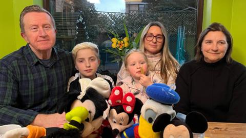 The Mead family - including Grandad Alan and grandmother Debbie with their daughter and her children. They are in a very bright green room. The children have Disney toys, including Mickey Mouse, Minnie Mouse and Donald Duck, sitting in front of them on the table.