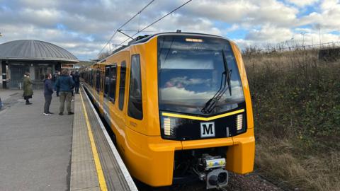 A yellow and black new style metro train at an outdoor station, with passengers waiting on the platform. Around building sits on the central platform between the two rail lines.