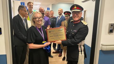 A man in a black miliitary-style uniform holds up a gold plaque alongside a woman in a black dress. Behind them are several people posing for the camera. 