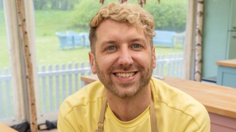 John Mincher, who has blonde hair and is wearing a yellow top, is in the Bake-off tent