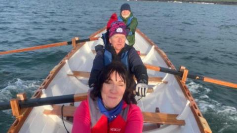 A group of people rowing a boat. Three of them can be seen - a woman with dark hair, a man in a wooly Scotland rugby had and another man in a green coat with a grey beard.