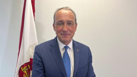 Jersey's chief minister is standing in front of a Jersey flag. He is wearing a navy blue blazer and light blue tie.