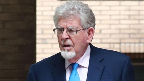 A close up of Rolf Harris. He has short white curly hair and is wearing a pair of glasses. He has white facial hair and is also wearing a navy jacket, light blue tie, and a white shirt.