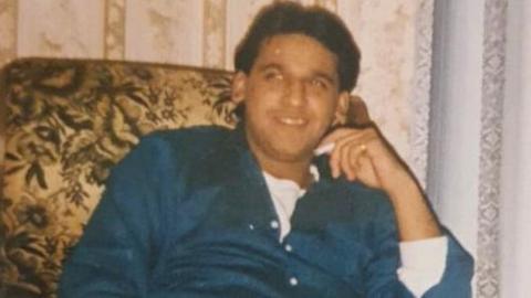 A photograph of Ethsham Ul-Haq Ghafoor smiling and holding a cigarette. His is sat down and wearing a blue shirt. 