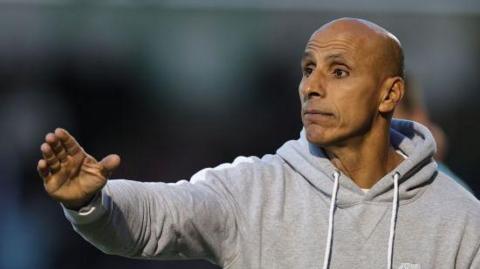 Dino Maamria pointing with his right hand, wearing a grey hoodie 