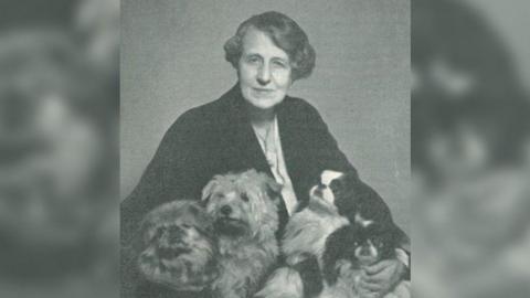 Black and white photo of Nina Duchess of Hamilton and Brandon holding four dogs in her arms while seated.