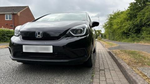 Black Honda Jazz parked a short distance from a kerb, with block paving between the pavement and the car. There is a hedge to the right of the picture and a modern two-storey house to the left.