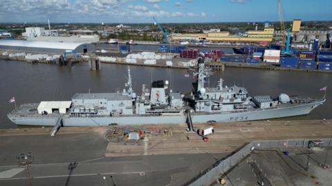 A grey warship is moored at a dock. Shipping containers, cranes and industrial warehouses are visible in the background.