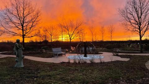 A garden with grass and paths running through it. It features a statue, a sculpture in the middle and a bench. There are also multiple leafless trees sitting under a bright orange sunset. 
