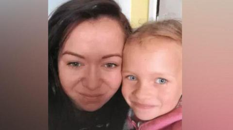 A woman with long dark hair and brown eyes is cuddled up next to a young girl with blonde hair and blue eyes wearing a pink top.