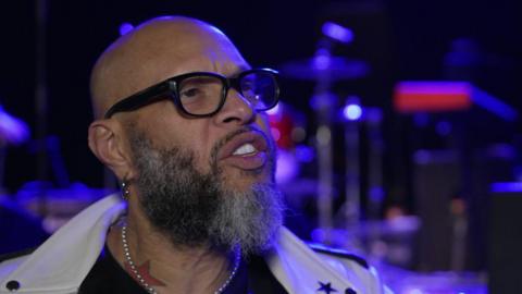 Frank Ferrer gives a TV interview inside a music venue. He is bald with a beard and wears a white leather jacket and jewellery.