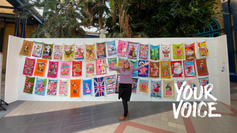 Large display of colourful tea towel artworks hung on lines, with one person standing in front of the installation.