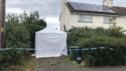 A white forensics tent outside a house. Blue and white police tape can also be seen, cordoning off the property. 