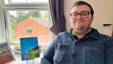 A man with brown hair and facial stubble. He is wearing black glasses and a blue and white top. On a white table behind him are two small paintings on small brown easels in front of a window. One has a blue lake next to grass, trees and a blue sky. The other is blue and orange water below a starry night sky.