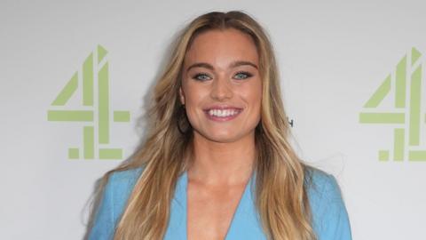 The image features Jodie Ounsley standing in front of a backdrop that displays the Channel 4 logo alongside the Paris 2024 Paralympic Games logo. She is dressed in a light blue suit, consisting of a blazer with a deep V-neckline and matching trousers. Her long, wavy hair cascades over her shoulders, and she is smiling widely, showing her teeth.